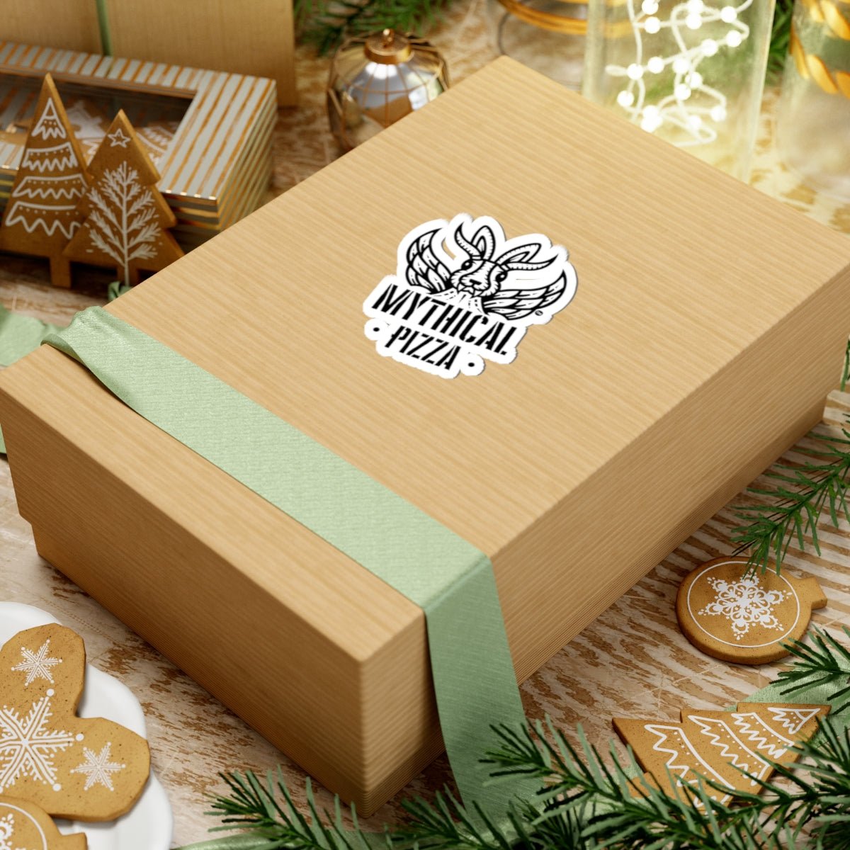 A square tan gift box wrapped with a sage green ribbon with a "Mythical Pizza" sticker sits among Christmas decorations.