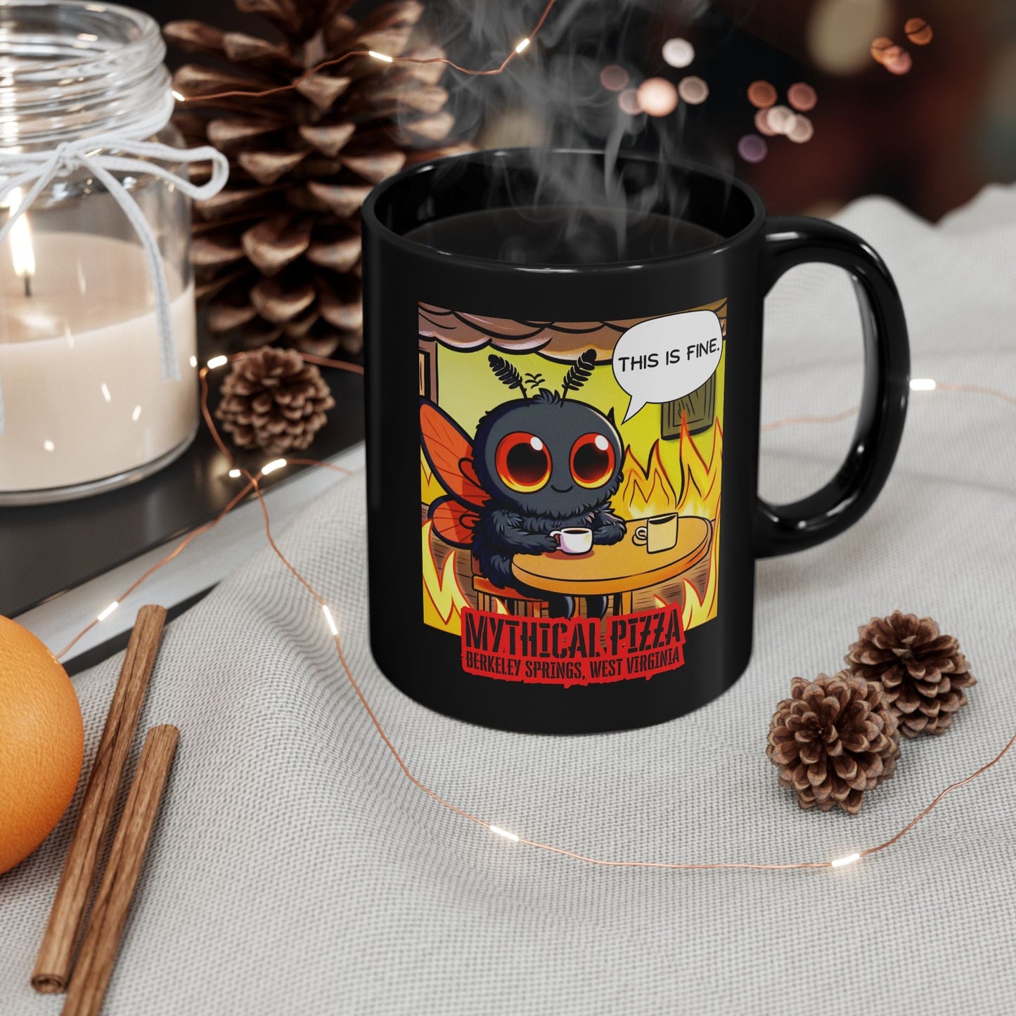 Close-up of a black mug with a cartoon mothman character drinking coffee amidst flames, resting on a table with the words "Mythical Pizza, Berkeley Springs, West Virginia" printed on it, steam rising from the mug's contents. The mug is surrounded by pine cones, fairy lights, an orange, cinnamon sticks, and a candle in a glass jar on a textured gray surface.